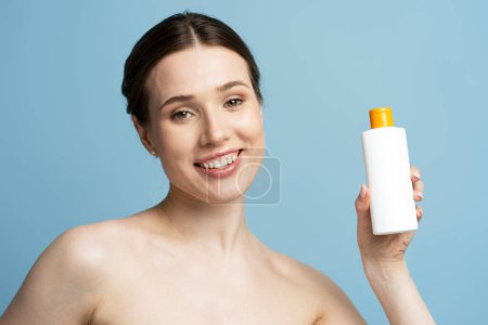 Foto de Atractiva mujer sonriente sosteniendo botella de champú mirando a la cámara aislada sobre fondo azul, concepto de cuidado del cabello. Burla. - Imagen libre de derechos