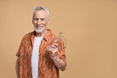 Foto de Hombre mayor barbudo con estilo de pelo gris con camisa de verano naranja, sosteniendo refrescante limonada, sonriendo una sonrisa dentada mirando a la cámara, aislado sobre un fondo beige. Vacaciones. Vacaciones. Copiar espacio - Imagen libre de derechos