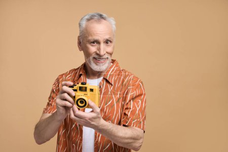 Téléchargez les photos : Charmant homme âgé caucasien élégant, voyageur touristique en chemise décontractée orange, tenant caméra jaune moderne, souriant sur fond isolé beige. Personnes, tourisme, voyage, vacances d'été concept - en image libre de droit
