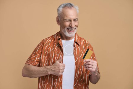 Téléchargez les photos : Homme âgé caucasien, retraité retraité voyageur touristique positif, en chemise orange, sourires et clins d'oeil, montrant un pouce vers le haut en regardant la caméra, posant avec une carte de crédit bancaire isolé sur fond beige - en image libre de droit