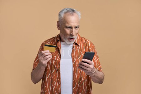 Téléchargez les photos : Incroyable beau souriant homme âgé caucasien des années 60, tenant une carte de crédit en plastique doré, en utilisant une application mobile, shopping en ligne, paris sportifs isolé fond bleu. Les gens. La finance. Concept technologique - en image libre de droit