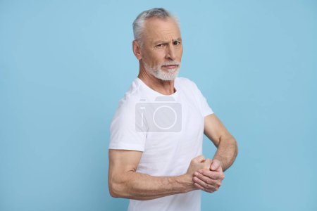 Téléchargez les photos : Attrayant homme âgé caucasien musclé, beau et imposant adulte en t-shirt blanc, montrant les muscles biceps, regardant la caméra, sur fond bleu isolé. Des retraités actifs. Modes de vie sains - en image libre de droit