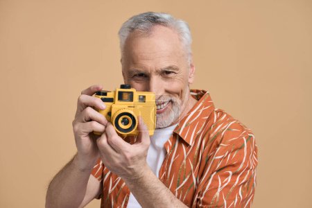 Téléchargez les photos : Charmant homme âgé caucasien, touriste voyageur en chemise décontractée orange, tenant un appareil photo jaune vif, souriant largement tout en prenant des photos, isolé sur fond beige. Les gens et le concept d'été - en image libre de droit