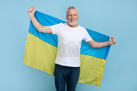 Téléchargez les photos : Beau conscient caucasien cheveux gris barbu senior homme 60, volontaire, patriotique citoyen sourit en regardant caméra, portant sur ses épaules le drapeau de l'Ukraine, isolé sur fond bleu - en image libre de droit