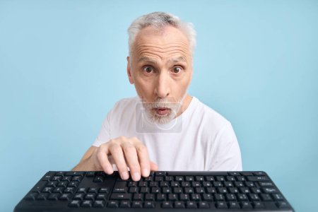 Foto de Sorprendido hombre mayor de 60 años agarrado de la mano en el teclado de la computadora PC, expresando sorpresa, haciendo girar los ojos y posando con la boca abierta, sobre un fondo azul aislado. Gente. Tecnología. Punto de vista portátil - Imagen libre de derechos