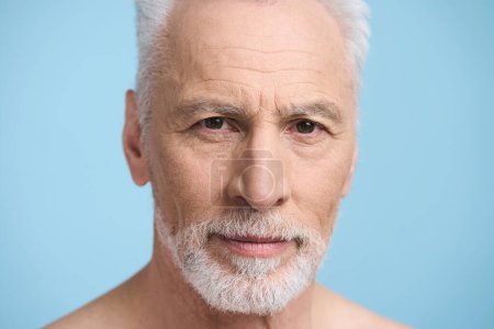 Photo for Close-up studio portrait. Face of Caucasian handsome gray haired bearded senior man 60s, looking at camera, isolated on blue background. Healthy lifestyle. Male beauty. Barbershop. Skin and body care - Royalty Free Image