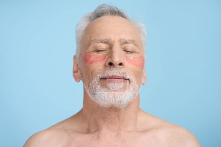 Photo for Close-up portrait of Caucasian handsome relaxed senior man 60s, wearing pink collagen hydrogel patches under eyes to soothe redness and puffiness, standing bare shoulders over isolated blue background - Royalty Free Image