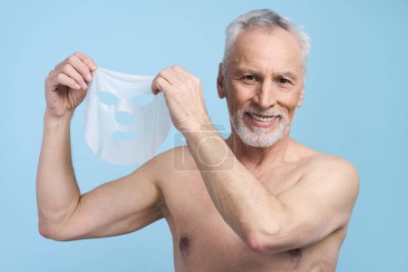 Téléchargez les photos : Bel homme blanc âgé, 60 ans debout avec torse nu sur fond bleu isolé, souriant à la caméra, tenant un masque de tissu pour hydrater nourrir et purifier la peau du visage - en image libre de droit
