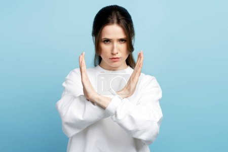 Photo for Strict bossy woman with brown hair in white sweater showing x sign with hands, there is no way more, hopelessness. Indoor studio shot isolated on blue background - Royalty Free Image