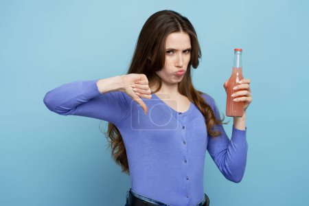 Photo for Portrait of unhappy sad woman in blue shirt making finger down and holding beverage. Studio shot isolated on blue background - Royalty Free Image