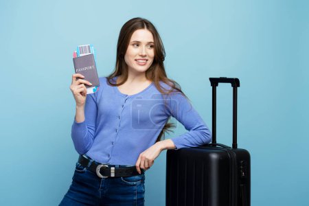 Photo for Cheerful caucasian young happy woman, passenger tourist traveler going for weekend getaway, holding passport with flight ticket, smiling cutely, posing with suitcase on blue background - Royalty Free Image