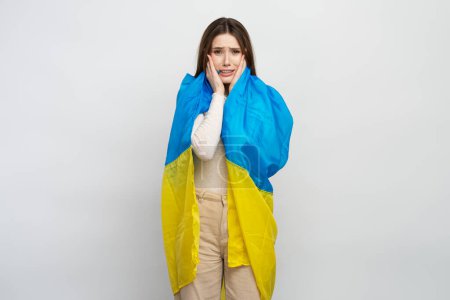 Téléchargez les photos : Belle jeune femme ukrainienne, enveloppée dans le drapeau bleu et jaune de l'Ukraine, pleurant isolé sur fond blanc, ressentant la douleur à cause de la guerre dans son pays. Arrêtez la guerre - en image libre de droit