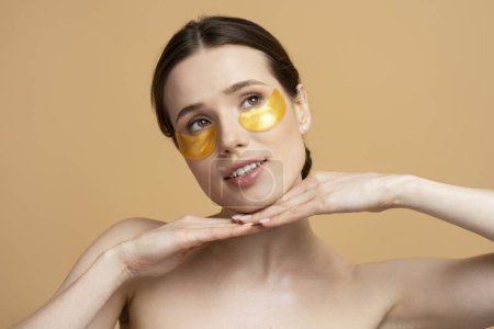 Téléchargez les photos : Portrait rapproché de belle femme caucasienne souriante utilisant des patchs dorés sous les yeux isolés sur fond beige. Soins de la peau, masque facial, concept de beauté naturelle - en image libre de droit