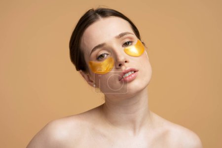 Photo for Closeup beauty portrait of young caucasian beautiful woman with golden hydrogel anti puffiness patches under eyes, holding her hands under face, smiling looking at camera, isolated beige background - Royalty Free Image