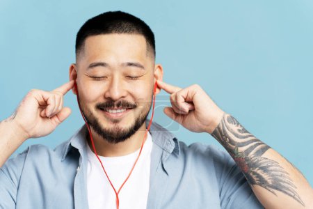 Téléchargez les photos : Homme coréen attrayant écoutant de la musique dans les écouteurs, les yeux fermés avec des tatouages élégants sur la main isolé sur fond bleu. Homme relaxant dans des vêtements décontractés élégants, concept de technologie moderne, profiter - en image libre de droit