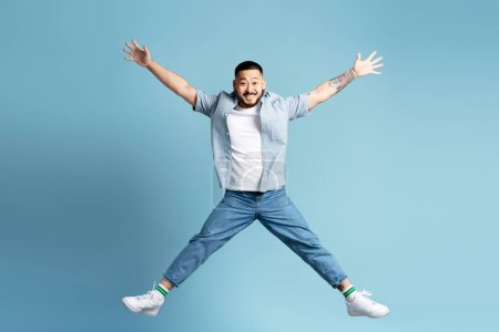 Photo for Funny smiling Asian man with stylish tattoos on his hands jumping isolated on blue background. Attractive male looking at camera, happy - Royalty Free Image