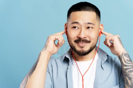 Téléchargez les photos : Gros plan de bel homme asiatique écoutant de la musique, portant des écouteurs, regardant loin isolé sur fond bleu. Beau mâle dans des vêtements décontractés élégants avec des tatouages élégants. Concept technologique - en image libre de droit