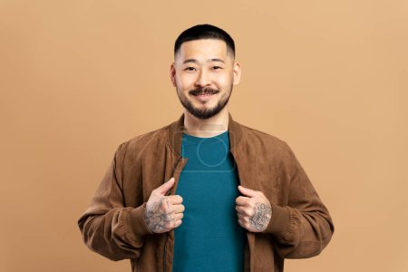Téléchargez les photos : Portrait d'un coréen souriant vêtu d'une veste brune, isolé sur fond beige. Homme d'affaires attrayant avec des tatouages élégants sur ses bras - en image libre de droit