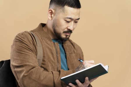 Foto de Estudiante asiática seria y ofensiva escribiendo en cuaderno con bolsa aislada sobre fondo beige. Hombre atractivo en chaqueta marrón - Imagen libre de derechos