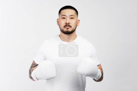 Téléchargez les photos : Portrait d'un homme asiatique sérieux portant des gants de boxe blancs isolés sur fond blanc. Entraînement des jeunes hommes, motivation, passe temps - en image libre de droit