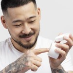 Closeup of Asian man with stylish tattooed hands picking cream with finger isolated on white background. Skin care concept after shower