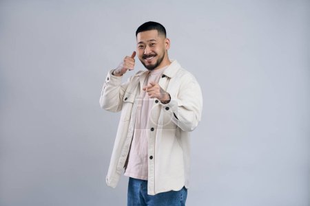 Photo for Portrait of handsome asian man in casual shirt standing with telephone hand gesture and pointing to camera, offering to contact by phone. Indoor studio shot isolated on grey background - Royalty Free Image