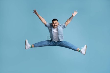 Foto de Retrato completo de hombre asiático inspirado positivo saltando en el aire. Estudio interior plano aislado sobre fondo azul, espacio de copia vacío - Imagen libre de derechos