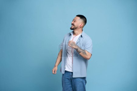 Foto de Hombre coreano feliz satisfecho con tatuaje en el brazo, usando auriculares disfrutando de música aislada escuchando canción favorita sobre fondo azul, espacio de copia. Tecnología, concepto de emociones positivas - Imagen libre de derechos