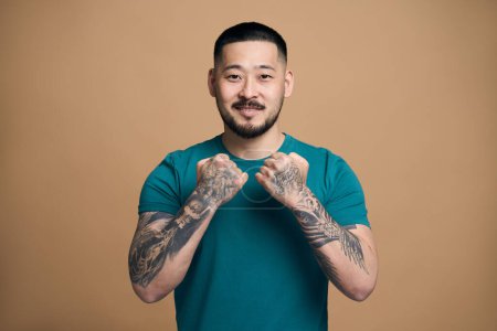 Foto de Retrato de un guapo hombre barbudo asiático con ropa de estilo casual mostrando brazos tatuados y posando con expresión facial positiva, mirando a la cámara con sonrisa. Aislado sobre fondo marrón - Imagen libre de derechos
