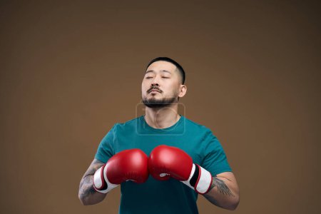 Photo for Motivated asian man in green t shirt and red boxing gloves standing over brown background with closed eyes and relaxing - Royalty Free Image