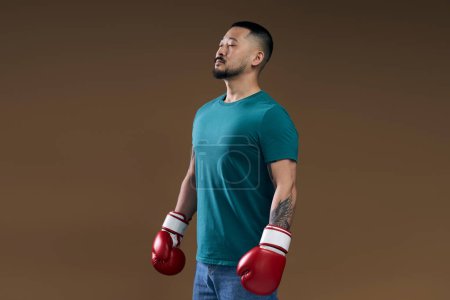 Photo for Confident young fit korean sportsman in green t shirt wearing red boxing gloves posing with closed eyes isolated over brown background - Royalty Free Image