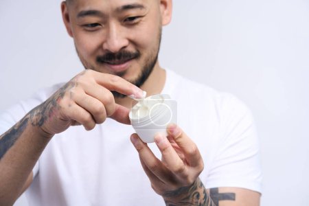 Photo for Cheerful asian man applying cream on face, holding moisturizer jar in hand, smiling while standing isolated at white background. Male skin care and beauty concept - Royalty Free Image