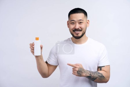Foto de Joven hombre asiático con camiseta blanca sosteniendo botella de champú sonriendo feliz y señalando con el dedo a los cosméticos. Tatuado chico recomendando gel para el cabello sobre fondo de pared blanco - Imagen libre de derechos