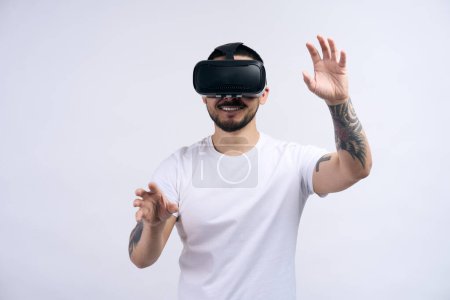 Téléchargez les photos : Homme asiatique excité portant t-shirt blanc dans des lunettes vr jouer à des jeux vidéo avec casque de réalité virtuelle, en essayant de toucher quelque chose avec la main isolé sur fond de mur blanc - en image libre de droit