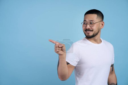 Photo for Smiling asian bearded man pointing to side copy space with one hand and showing blank wall for idea presentation. Indoor studio shot isolated on blue background - Royalty Free Image