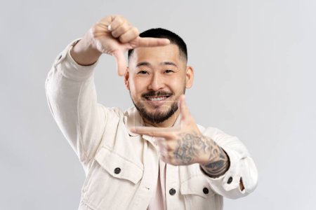 Foto de Retrato de confianza feliz asiático fotógrafo haciendo marco con las manos mirando cámara aislada sobre fondo blanco - Imagen libre de derechos