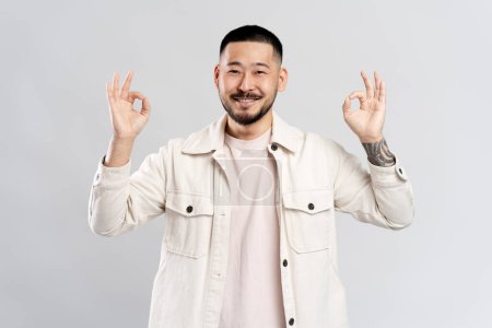 Téléchargez les photos : Attrayant souriant asiatique homme portant une chemise décontractée élégante montrant ok signe, regardant la caméra isolée sur fond gris. Concept des émotions positives - en image libre de droit