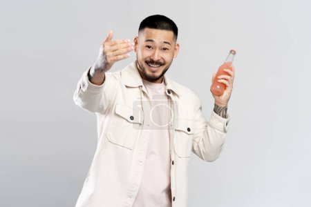 Téléchargez les photos : Amical souriant asiatique homme portant des vêtements décontractés, tenant bouteille avec boisson inviter à la fête, isolé sur fond blanc. Concept de style de vie positif - en image libre de droit
