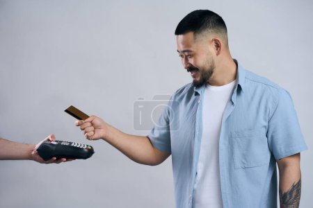 Foto de Atractivo sonriente hombre asiático sosteniendo tarjeta de crédito haciendo el pago utilizando la máquina de tarjeta de crédito, aislado sobre fondo gris. Compras, concepto de tecnología inalámbrica - Imagen libre de derechos