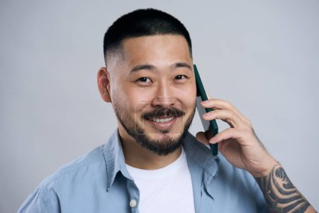 Foto de Retrato de sonriente hombre asiático confiado hablando en el teléfono móvil mirando a la cámara, aislado sobre fondo gris. Concepto tecnológico - Imagen libre de derechos