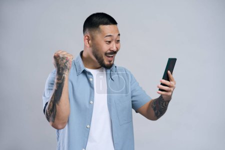 Téléchargez les photos : Homme asiatique excité tenant téléphone mobile, communication en ligne, ayant appel vidéo isolé sur fond gris. hipster coréen émotionnel en utilisant l'application mobile, jouer au jeu - en image libre de droit