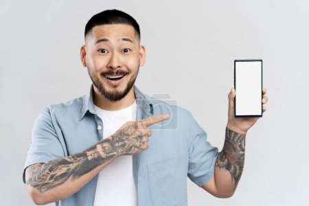 Téléchargez les photos : Homme asiatique sorti avec tatouage élégant tenant téléphone mobile faisant des achats en ligne pointant du doigt à l'écran vide, isolé sur fond blanc. Homme heureux moderne utilisant l'application mobile, commander de la nourriture. Maquette - en image libre de droit