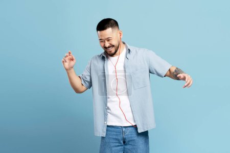 Foto de Atractivo hombre asiático feliz con un tatuaje elegante, con auriculares escuchando música, bailando aislado sobre fondo azul, espacio de copia. Tecnología, concepto de emociones positivas - Imagen libre de derechos