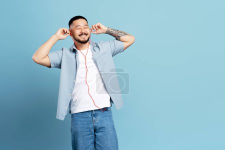 Téléchargez les photos : Beau homme asiatique heureux avec un tatouage élégant, portant des écouteurs écoutant de la musique isolée sur fond bleu, espace de copie. Technologie, concept des émotions positives - en image libre de droit