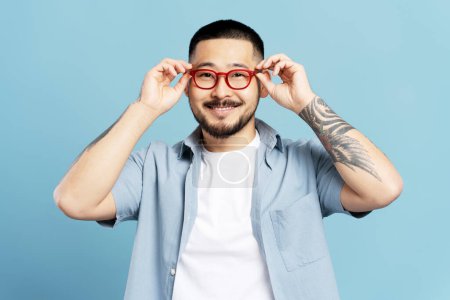 Foto de Retrato de hombre asiático guapo sonriente con tatuaje elegante aislado sobre fondo azul. Feliz hipster coreano con anteojos rojos mirando cámara. Concepto de visión - Imagen libre de derechos
