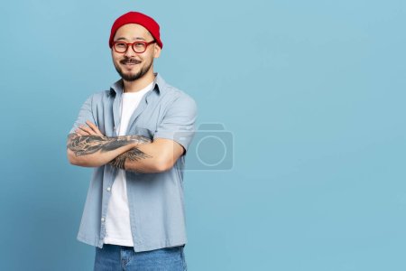 Foto de Guapo hombre coreano sonriente, trabajador exitoso con los brazos cruzados, tatuaje elegante aislado sobre fondo azul. Retrato asiático hipster con sombrero rojo, gafas mirando cámara. Negocios exitosos - Imagen libre de derechos