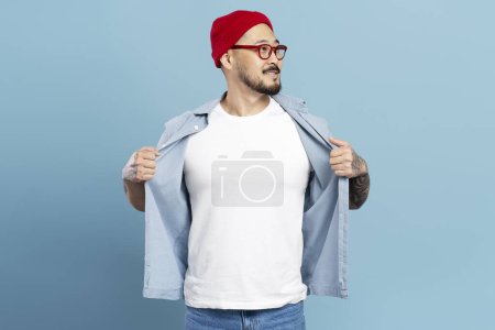 Foto de Sonriente hombre asiático seguro de sí mismo, hipster elegante con sombrero rojo, gafas, mostrando camiseta blanca con maqueta. Modelo coreano posando para fotos, aislado sobre fondo azul. Tienda, concepto de compras - Imagen libre de derechos