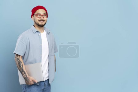 Photo for Portrait of smiling Korean freelancer with stylish tattoo, wearing red hat, eyeglasses looking at camera. Smart asian student holding laptop computer isolated on blue background, education concept - Royalty Free Image