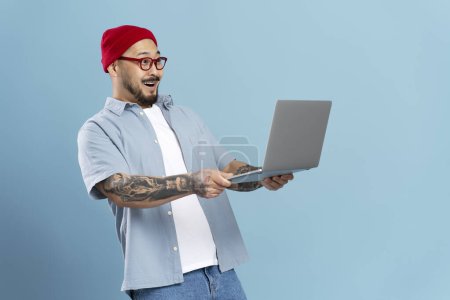 Téléchargez les photos : Homme asiatique élégant excité tenant ordinateur portable, regarder des vidéos avec connexion Internet haute vitesse 5g isolé sur fond bleu. Choqué hipster gars shopping en ligne avec des ventes. Technologie moderne - en image libre de droit