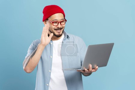 Téléchargez les photos : Souriant asiatique homme, pigiste tenant ordinateur portable, agitant la main, ayant appel vidéo - en image libre de droit
